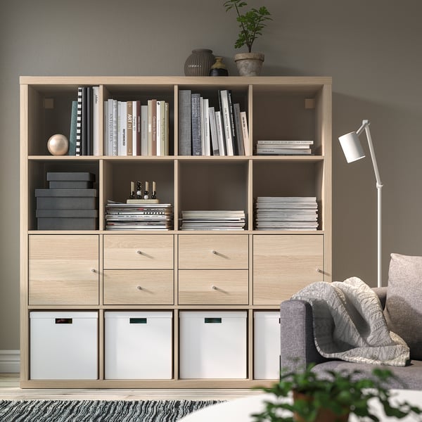 Wooden KALLAX shelf with books and decor, drawers, grey couch, white lamp.