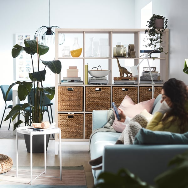 Person on blue sofa using phone, KALLAX shelving with plants, bright room.
