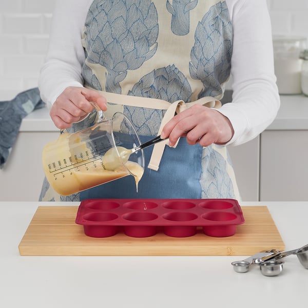 A person pours batter into a red silicone muffin mould set on a wooden board. The mould is for baking muffins, omelettes, or frozen desserts.