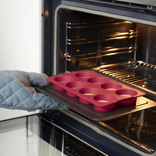 A hand, wearing a grey oven mitt, holds a red silicone muffin tray with eight cups, ready to be placed in an open oven.