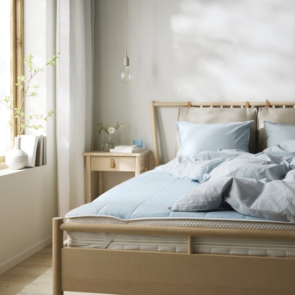 Cosy bed with light blue sheets on wooden frame, bedside table with books and flowers, white bulb hanging, light grey walls.