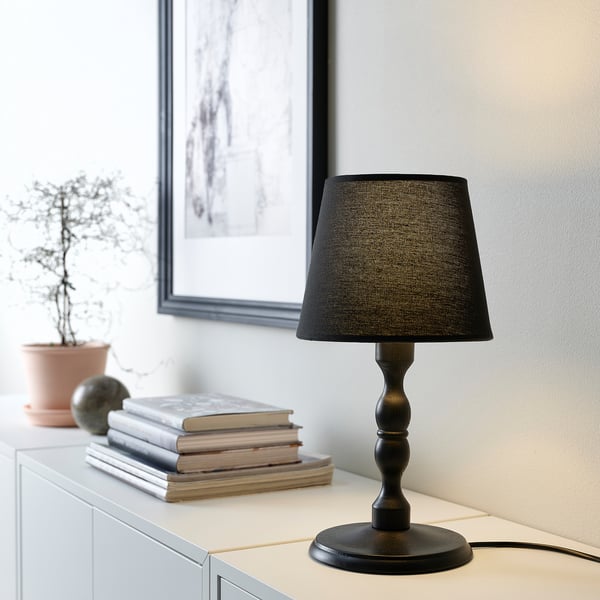 Black KINNAHULT lamp on dresser, shaped like candle holder, near books, plant, and frame.