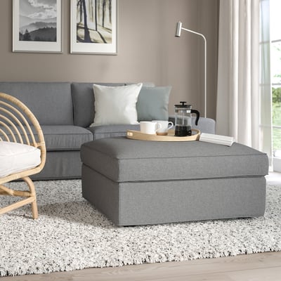 Modern living room: grey KIVIK ottoman, large, fabric-covered, near sofa & wicker chair. Provides extra seating, footrest, & hidden storage.