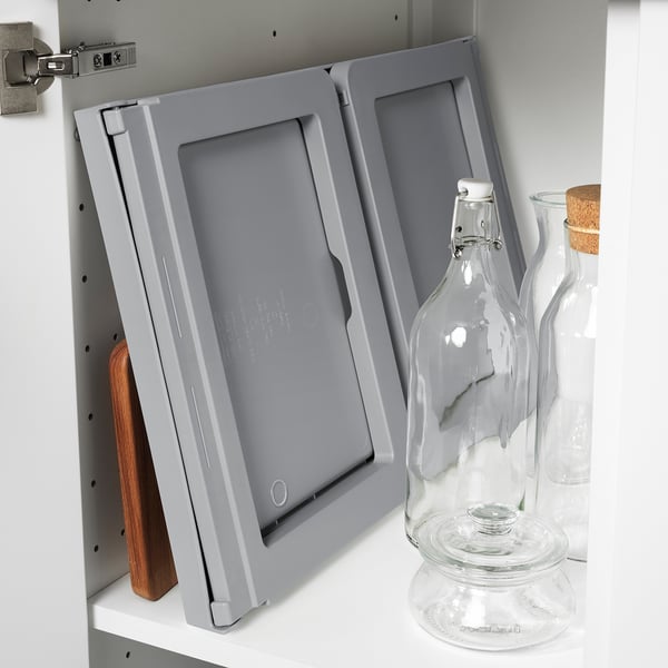 Grey KLIPSK bed tray stored vertically, leaning against cabinet, with glass jars beside it.