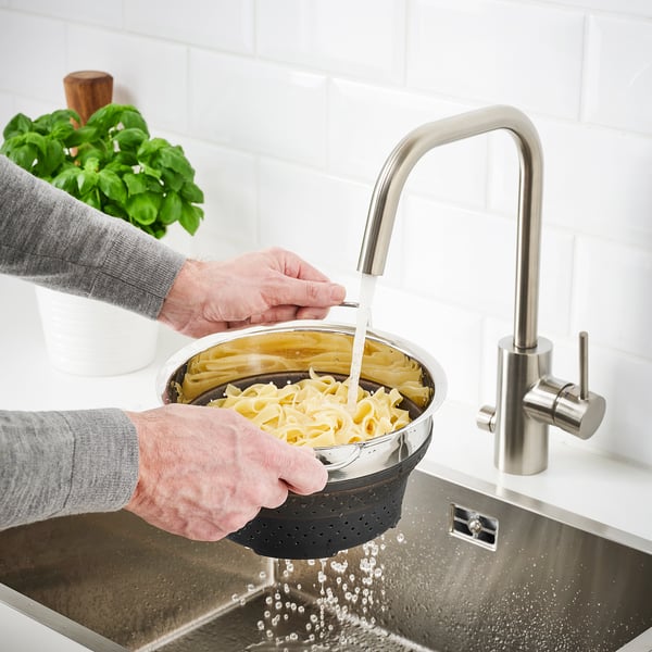Person rinsing cooked pasta in a black, foldable colander under a modern kitchen tap.