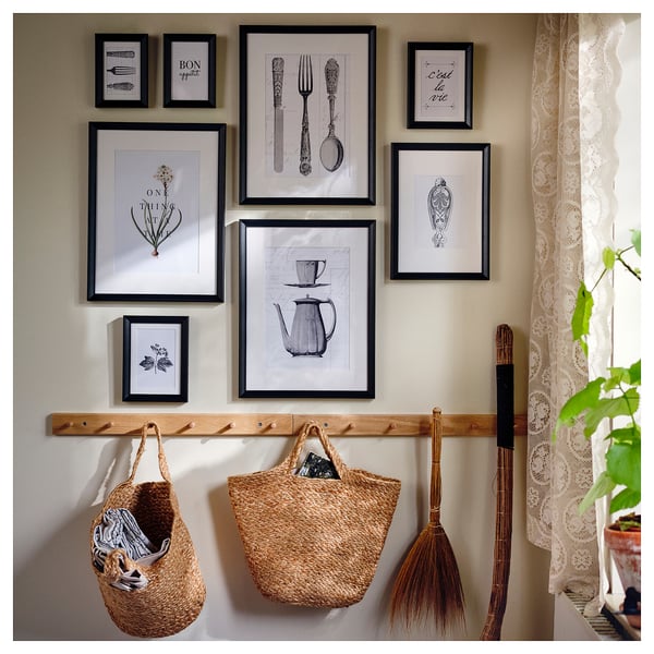 Gallery wall with several black-framed art pieces, hanging wicker baskets, and a broom. Lace curtain on the right.
