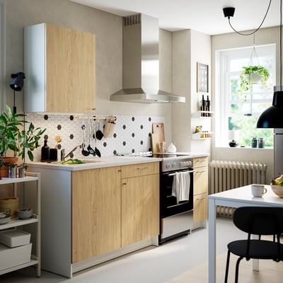 Modern kitchen with light wood cabinets, white tile backsplash, stainless appliances, black accents, green plants, and dining table.