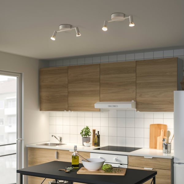 Modern kitchen with KRUSNATE ceiling light, featuring 2 adjustable spotlights. Wooden cabinets, white tiles, black island with bowl and avocado.