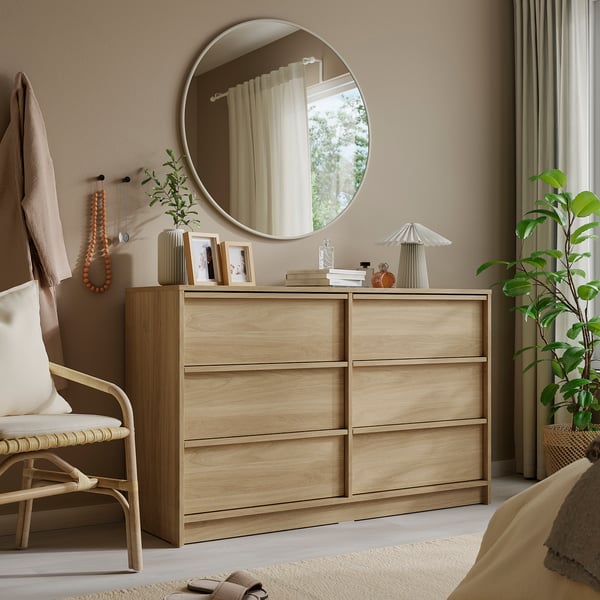 Round mirror with aluminium frame above wooden dresser. Bright room, six drawers, lamp, plant, wicker chair.