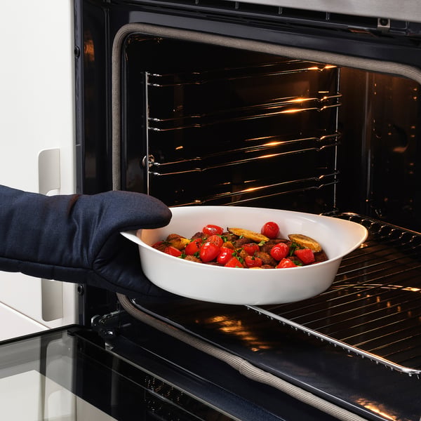 A hand wearing an oven mitt places a baking dish filled with food into an oven. The baking dish is used for both cooking and serving.
