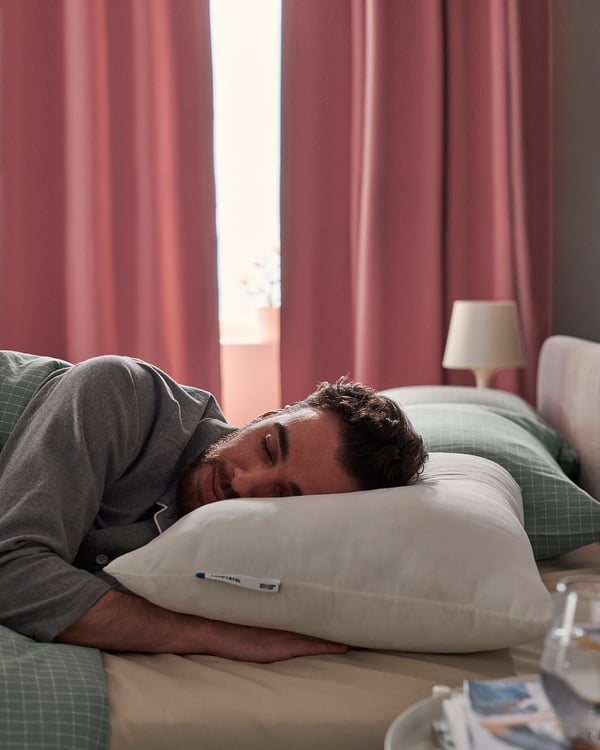 Person sleeps on a white LAPPTÅTEL pillow with grey shirt in a room with red curtains and green bedding.