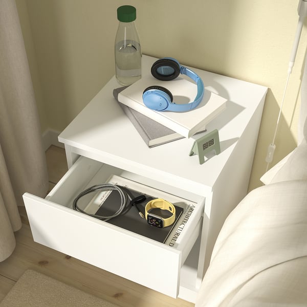 White bedside table with open drawer, closed compartment, and headphones on top.