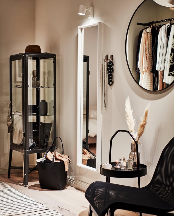 Black LINDBYN mirror with metal frame on wall, round shape, elegant design. Beside it, glass cabinet with items, black chair, and striped rug.