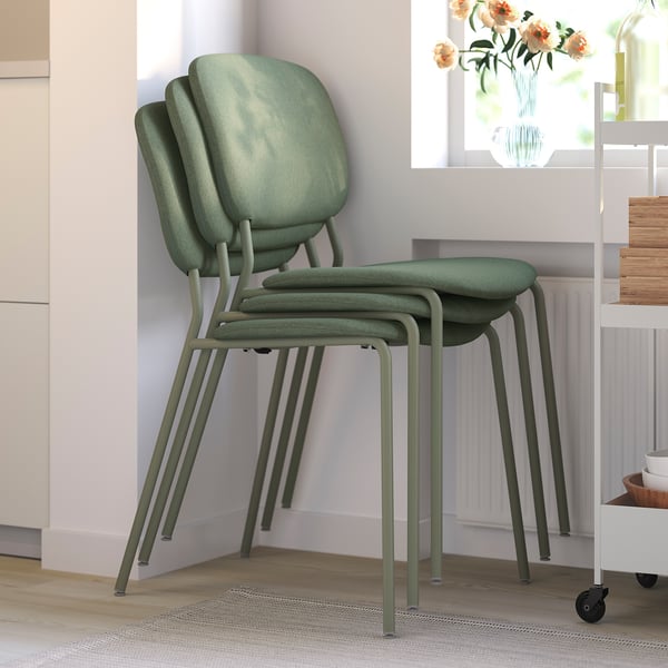 Three green stackable chairs, suitable for indoor use, neatly piled in a cosy room with light-colored walls and wooden flooring.