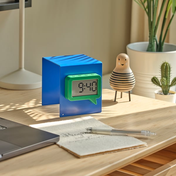 A blue cube clock with green speech bubble display at 9:40 on a wooden table with notebook, pen, laptop, and decorative items nearby.