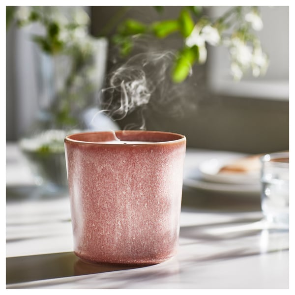 A pinkish-red LUGNARE scented candle emits smoke, sits on a table, featuring ceramic texture.
