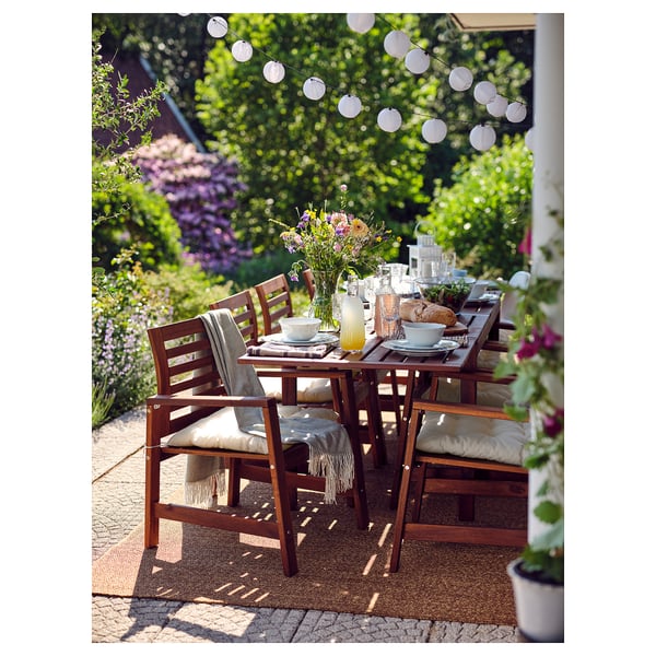 Outdoor dining setup with LYDERSHOLM brown wooden table, chairs, and rug. Decorated with dishes, drinks, and flowers.