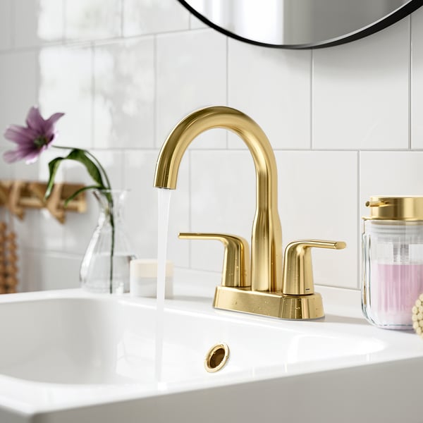 Gold tap pours water into white sink; set against tiled wall, with decorative vase and mirror nearby.