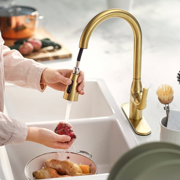 A person washes produce under a gold-colored tap with a pull-down spray head, showcasing its easy reach and cleaning functionality.