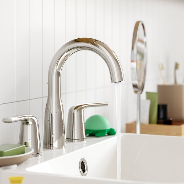 Modern bathroom tap with curved stainless steel design. Water flows gently from its sleek spout. Green soap dispenser and toothbrush holder nearby.