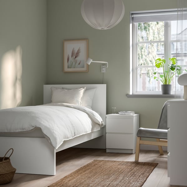 Minimalist bedroom with white MALM bed, sage walls, wall lamp, small bedside table, green vase, chair, rug, pendant light, city view window.