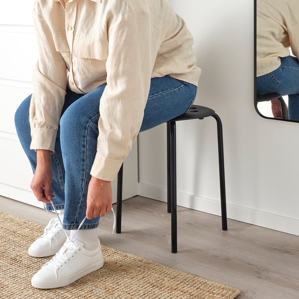 Person sitting on black MARIUS stool, tying white trainer, casual setting.
