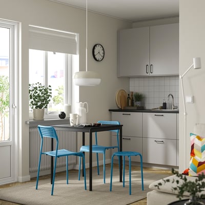 Small room with MARIUS blue plastic stools under a black table near a kitchen. Bright, modern design with a clock, plants, and light-colored walls.