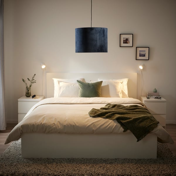 Modern bed with white bedding, green throw, blue lamp, wall photos, bedside table lamps.