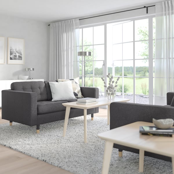 Modern living room with grey sofa, coffee table, and large windows. White pillows, cosy light.