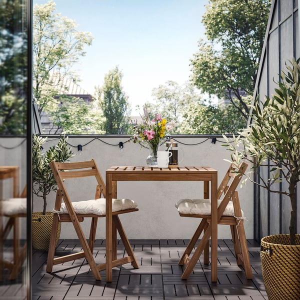 Small outdoor patio with wooden table, white cushioned chairs, vase of flowers, and string lights.