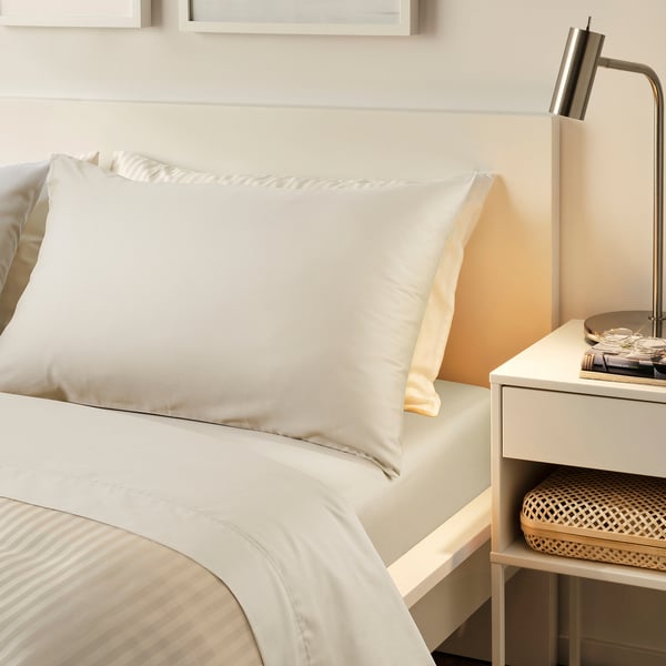 Elegant cream NATTJASMIN bed setup. Smooth silky sheets on large bed, bedside table, lamp, books.