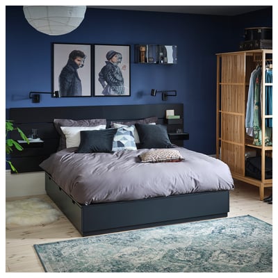 Modern bedroom with dark blue walls, grey bedding, wooden floor, and a black headboard. A wooden clothes rack with hangers and a patterned rug.