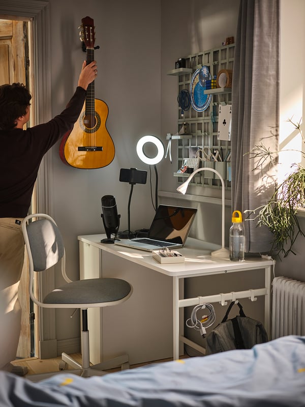 A person tunes a guitar beside a well-equipped desk with a laptop, microphone, ring light, and a white chair.