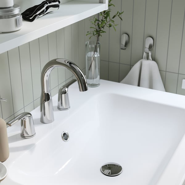 Modern chrome tap on white sink. Sleek, curved design with pop-up drain. Sits on stylish counter next to plant.