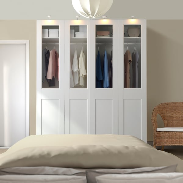 Modern white PAX wardrobe with glass doors showcasing clothes, standing tall beside a bed, featuring sleek design and soft lighting.