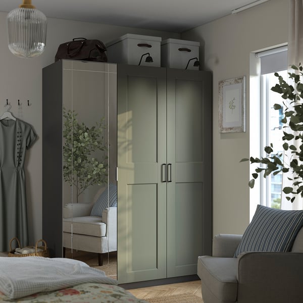 Grey-green PAX wardrobe with mirrored doors, top storage. Grey armchair by window with art. Cosy bed with grey sheets and floral pillows.