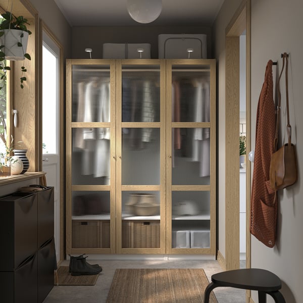 Large wooden PAX closet with glass doors occupies a room corner, taller than adjacent furniture, featuring organised storage spaces.