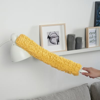 A person uses a PEPPRIG microfibre duster to clean dust from a wall shelf. The dusters cover is yellow, effective for large areas.