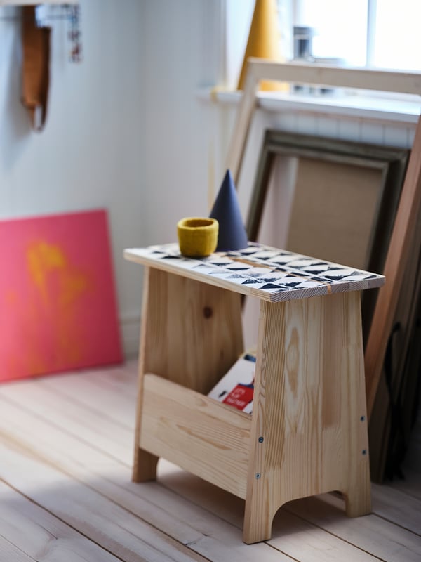 Light wooden stool with black and white tiled top, small yellow cup, blue cone, colourful artwork, natural light, minimalist design.