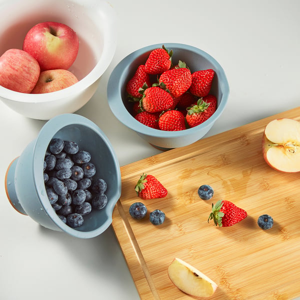 Three bowls filled with apples, strawberries, and blueberries. One bowl tilted on a cutting board with scattered fruit.