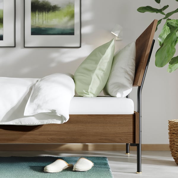 Bedroom scene with RÅDMANSÖ bed, green pillows, white blankets, wooden headboard, slatted base, and house slippers on colourful mat.