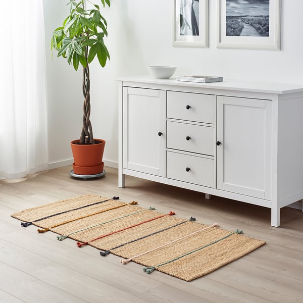 RAKLEV jute & wool rug with colourful fringes on wooden floor, between plant & sideboard.