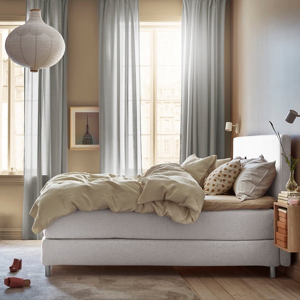 Bedroom with beige bedding, white bed, large windows with curtains, wall art, bedside table with books and lamp, large hanging white lamp.