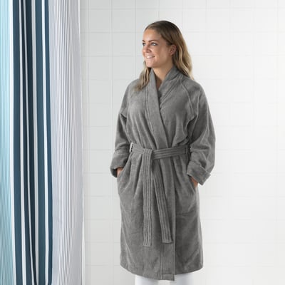 Person wearing grey ROCKÅN bathrobe with adjustable belt, side pockets, and cuffed sleeves, standing against tiled wall.