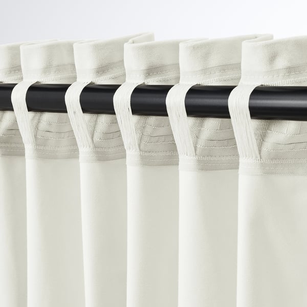 Off-white SANELA curtains hang elegantly with pleats. The black rod and tabs are visible, showing multi-way hanging options for versatile use.