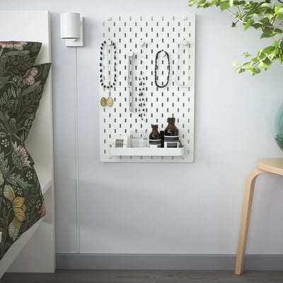 SKÅDIS pegboard with jewellery, bottles, clock. Beside bed with floral pillows and chair.