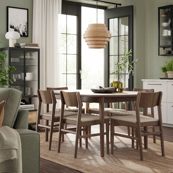 Modern dining room with SKANSNÄS dark wood table, four chairs, light fixture, and large windows. Green walls, white curtains, plants, and artwork.