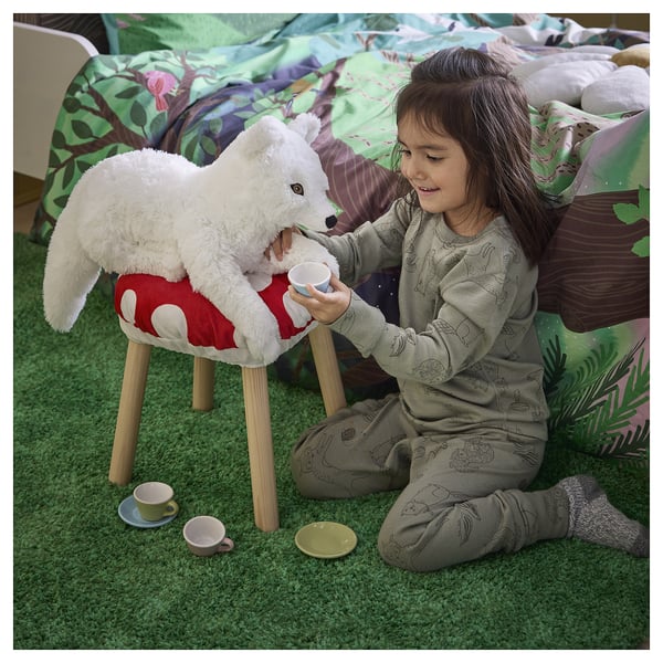Child plays tea with white fox stuffed animal on red stool. Green carpet, cups, saucer. Girl wears pyjamas, smiles.