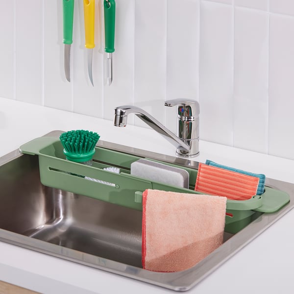 Stainless steel sink with green SKOLÄST organiser holding brush, sponges, and cloths near chrome tap.