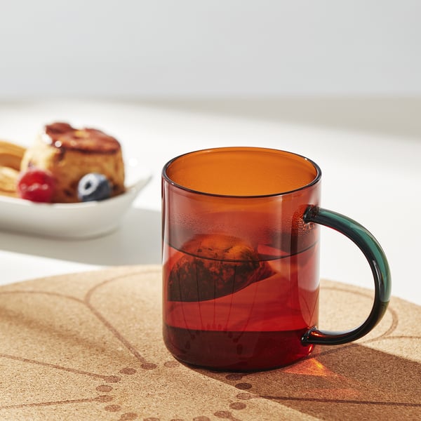 A red glass mug with a green handle sits on a cork mat, showcasing its comfortable grip and stylish design.
